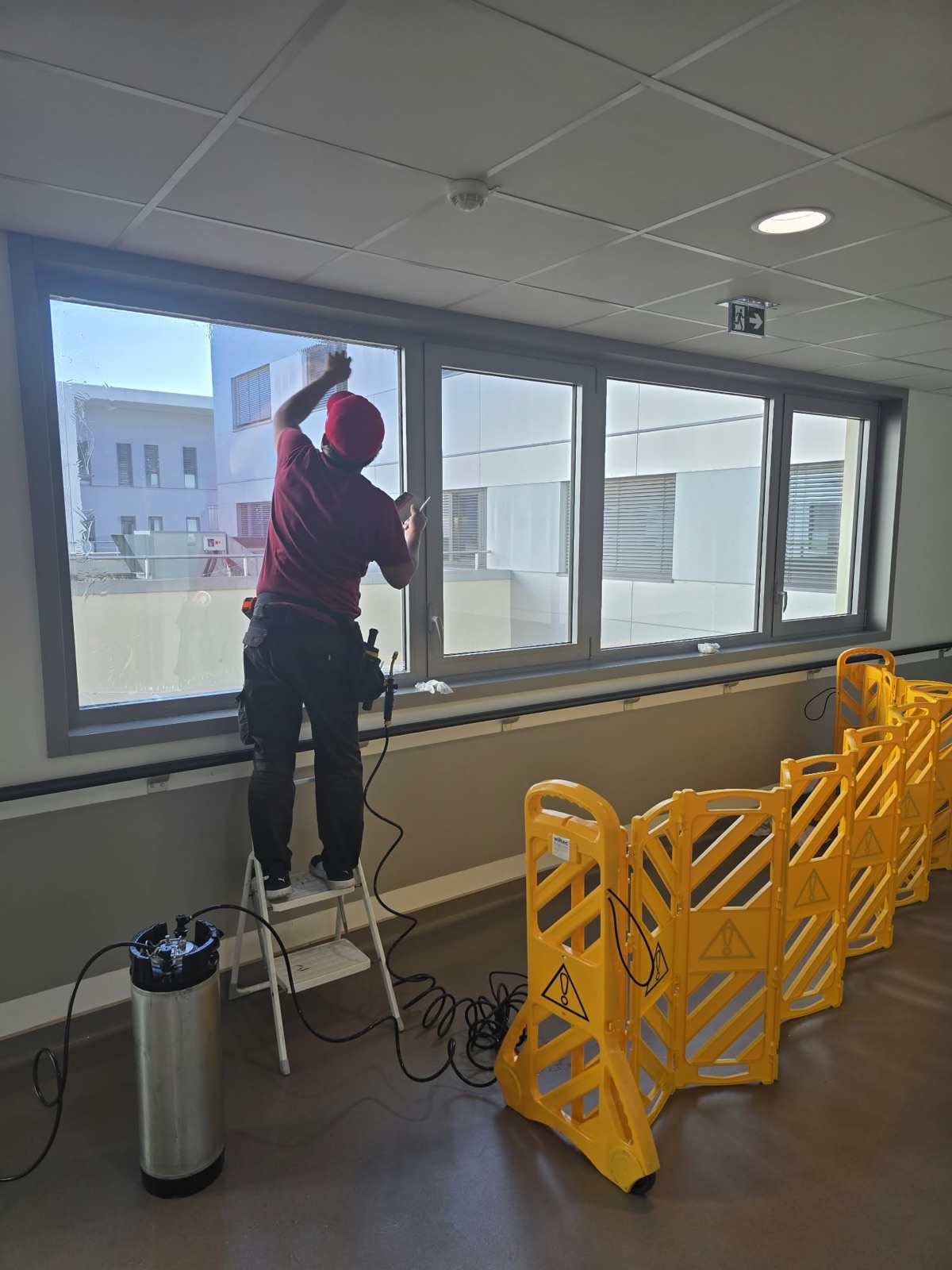 Technicien DFP France sur escabeau appliquant le film solaire sur une fenêtre de couloir de la Clinique Saint-Jean l'Ermitage, pulvérisateur au sol et barrières de balisage